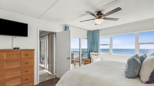 Ein Schlafzimmer mit einem großen Bett, einem Deckenventilator, einem Fernseher und Meerblick durch große Fenster.
