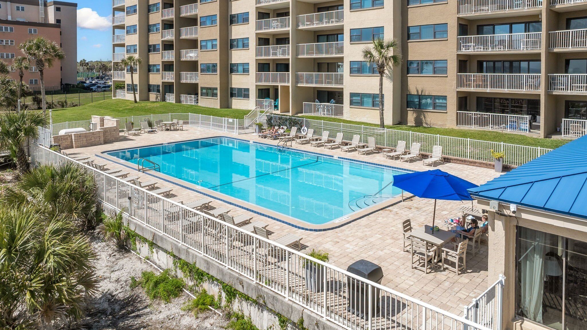 A swimming pool with lounge chairs and umbrellas, surrounded by a white fence and palm trees.