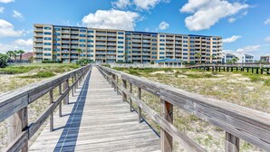 Une promenade en bois mène à un hôtel à plusieurs étages doté de balcons, situé sur une plage de sable où la végétation est clairsemée.