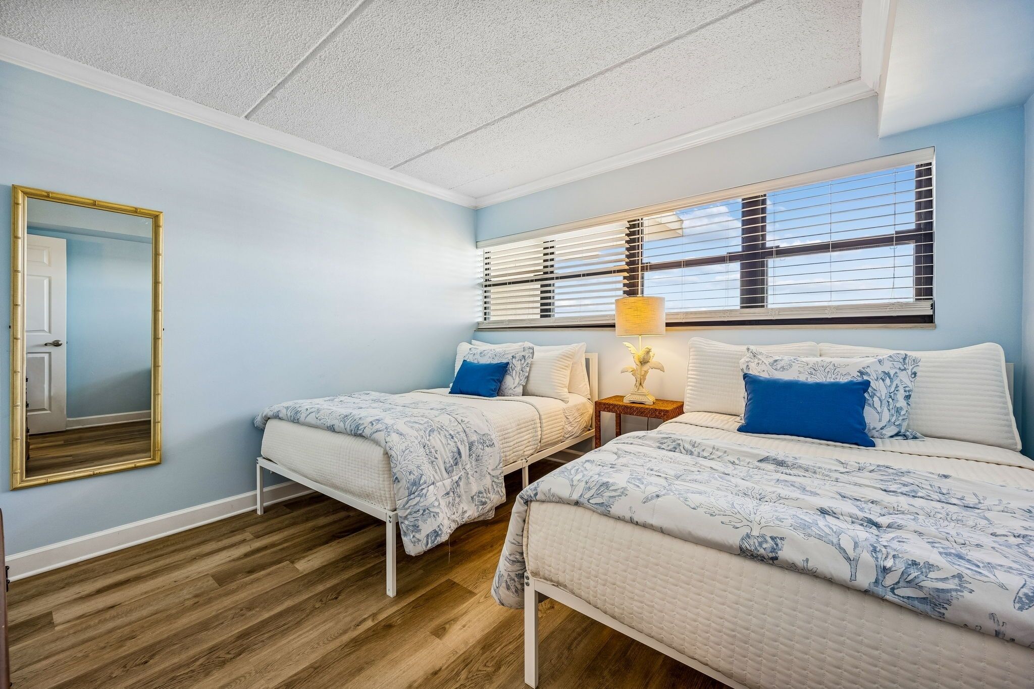 A bedroom with two beds, a large mirror, a window with blinds, and a bedside table with a lamp.