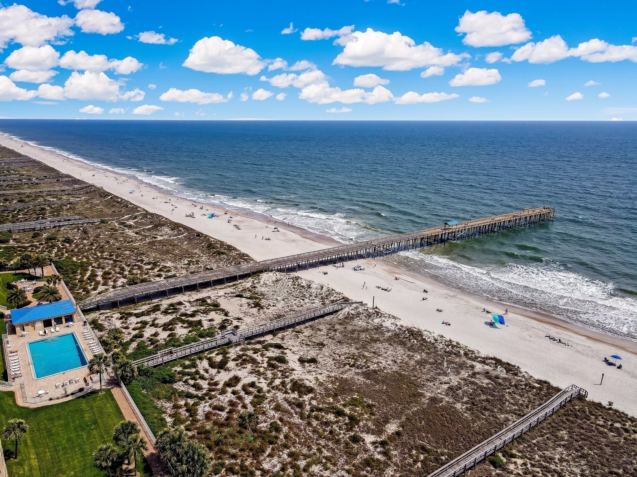 An aerial view of a beach with a long pier extending into the ocean, a swimming pool, and a landscaped area with palm trees.