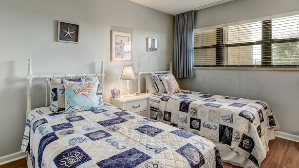 A bedroom with two beds, a nightstand, a lamp, and a window with blinds. - Convenience Direct Oceanfront Condo with Private Balcony by RedAwning (Fernandina Beach)