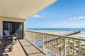 A balcony with a view of the beach and ocean, featuring a deck with chairs and a pool area. - Upper Unit Condo Overlooking Both the Ocean Pier and Swimming Pool by RedAwning (Fernandina Beach)