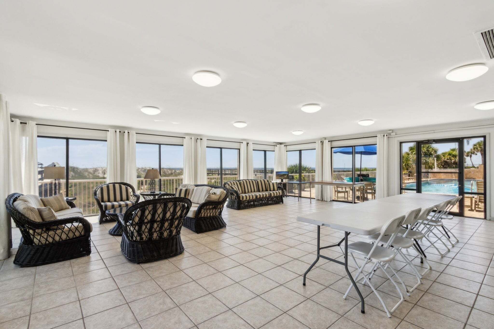 A spacious indoor area with large windows, wicker furniture, a long table, and a view of a pool and landscape.