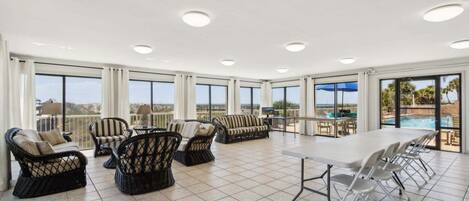 A spacious indoor area with large windows, wicker furniture, a long table, and a view of a pool and landscape.