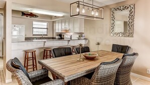 A dining area with a wooden table and wicker chairs, a hanging light fixture, and a decorative mirror on the wall. - Comfy Upper Unit Condo to Enjoy the Beach or the Fishing by RedAwning (Fernandina Beach)