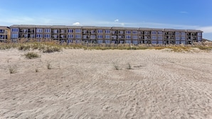 Um hotel de vários andares com telhado plano e amplas janelas, localizado numa praia arenosa com vegetação escassa.