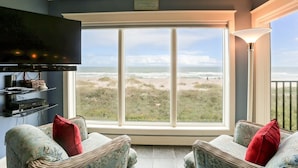 Sala de estar com uma grande janela com vista para a praia, uma TV de tela plana na parede e um abajur ao lado da janela.