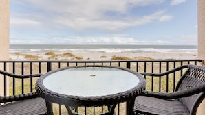 Varanda com mesa de vidro e cadeiras de vime, com vista para uma praia com ondas e dunas.