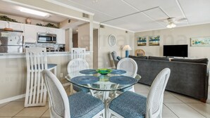 A modern living room with a glass-top dining table, white wicker chairs, a grey sofa, and a flat-screen TV.