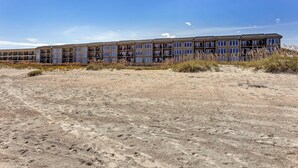 A multi-story hotel with a flat roof and large windows, situated on a sandy beach with sparse vegetation.