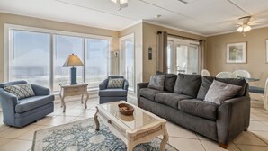 A spacious living room with a gray sofa, two blue armchairs, a glass coffee table, and a ceiling fan.
