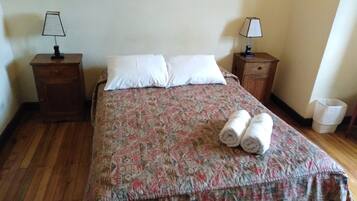 Cama bem arrumada com colcha estampada, dois travesseiros e duas toalhas enroladas. Há duas mesas de cabeceira com abajures e uma lixeira.