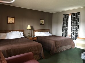 Quarto de hotel com duas camas, uma mesa de cabeceira com abajur, uma cadeira e uma janela com cortinas de bolinhas.
