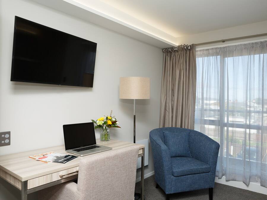 Ein Hotelzimmer mit flachbildfernseher an der Wand, einem Schreibtisch mit Laptop, einem Stuhl, einer Lampe und einem Fenster mit durchsichtigen Vorhängen.