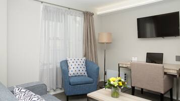 A modern hotel room with a sofa, a blue armchair, a desk with a laptop, a flat-screen TV, and a vase with flowers.