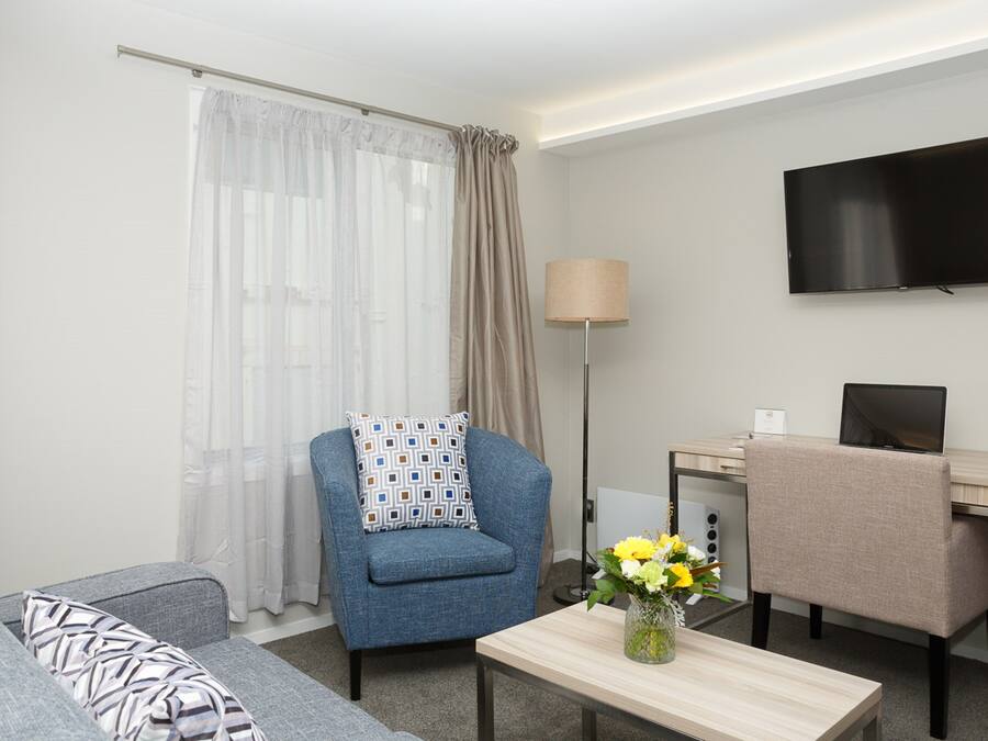 A modern hotel room with a sofa, a blue armchair, a desk with a laptop, a flat-screen TV, and a vase with flowers.