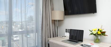 Une chambre d’hôtel équipée d’un bureau, d’un ordinateur portable, d’une lampe, d’un téléviseur à écran plat, d’un vase avec des fleurs et d’une fenêtre avec des rideaux.