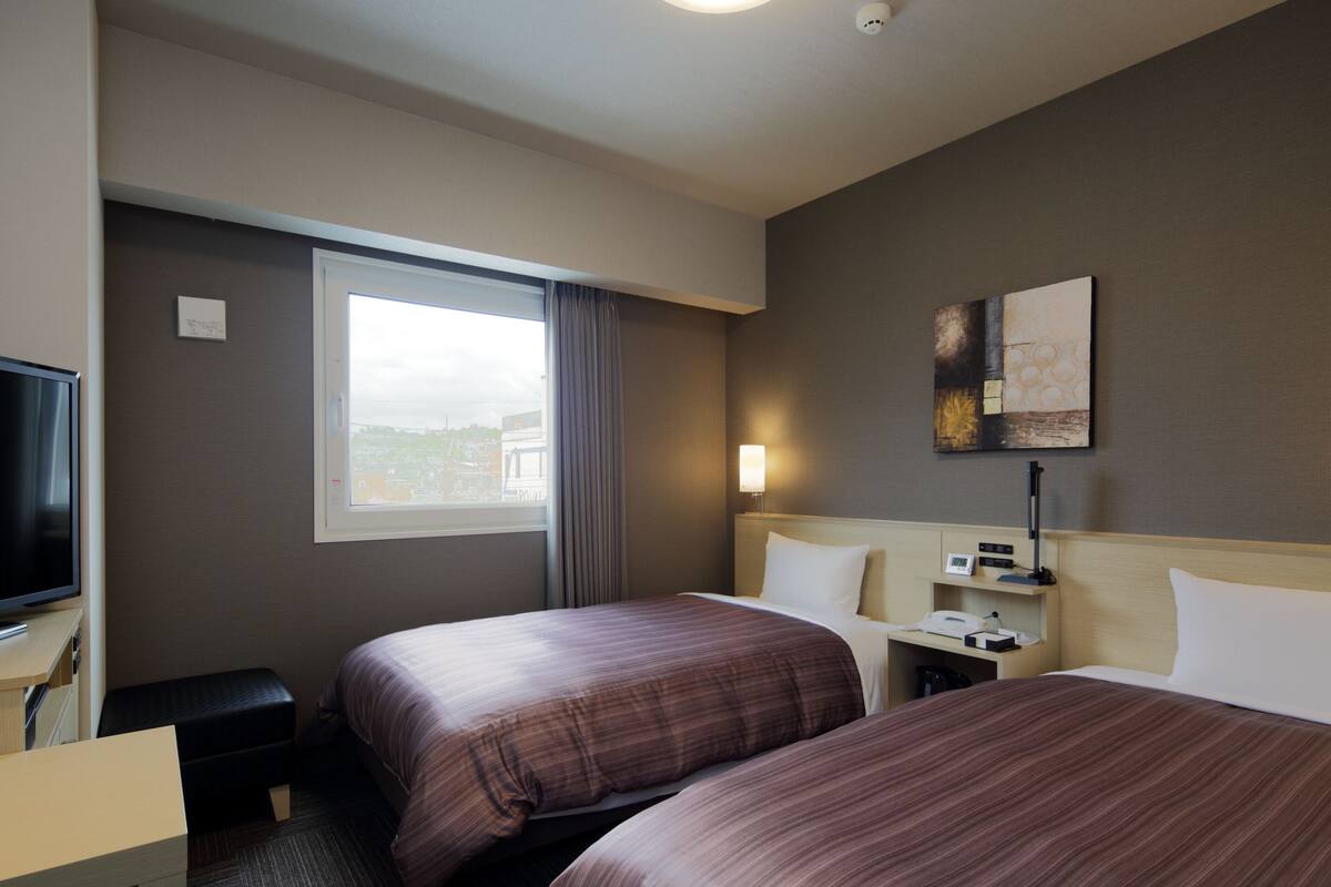 Habitación de hotel con dos camas, televisión, escritorio, lámpara y ventana con cortinas.
