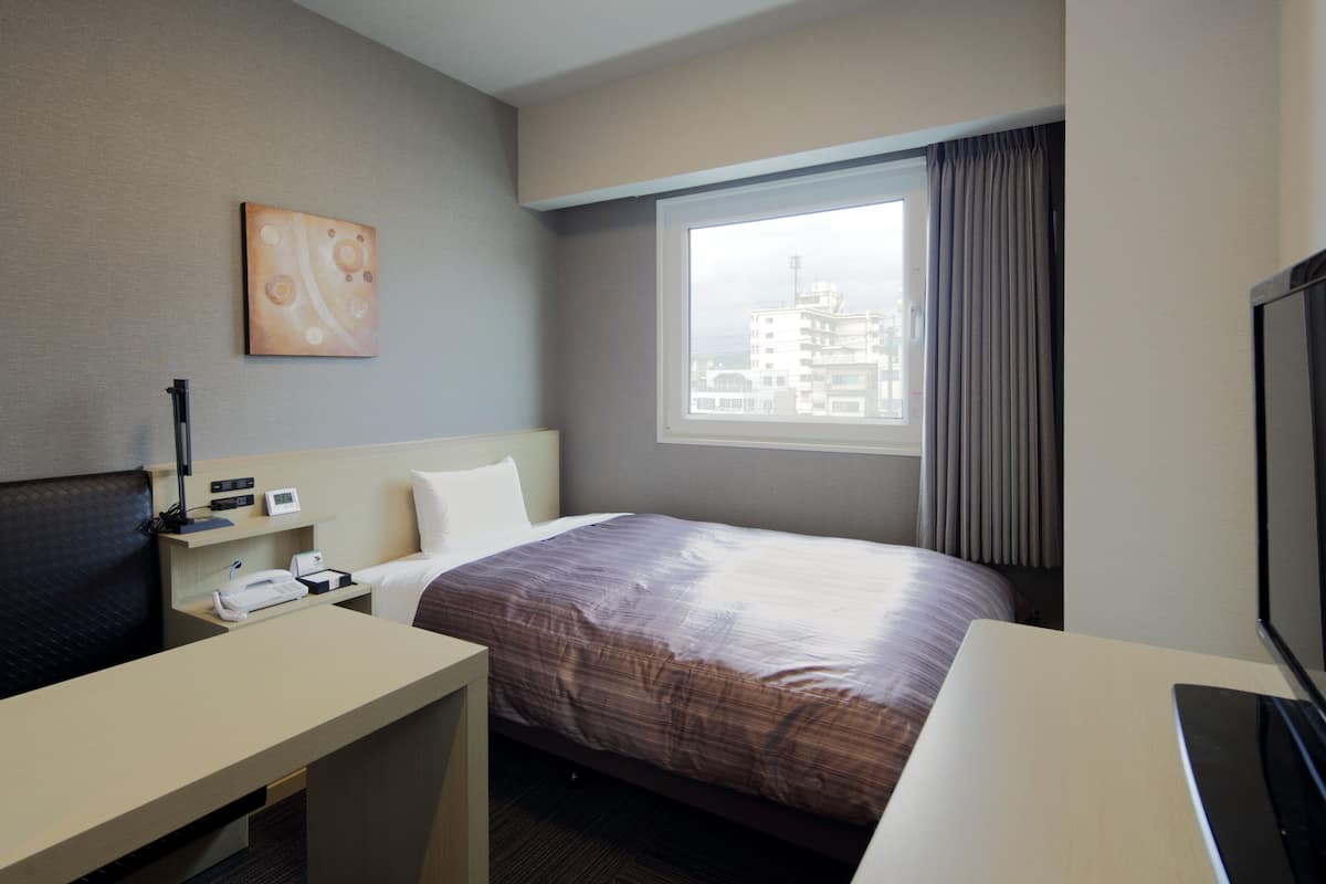 Habitación de hotel con cama, escritorio, silla, televisor, ventana con vista a la ciudad y un cuadro en la pared.