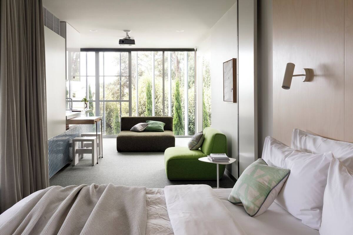 A modern hotel room with a large bed, a green armchair, a small table, and a view of greenery through large windows.