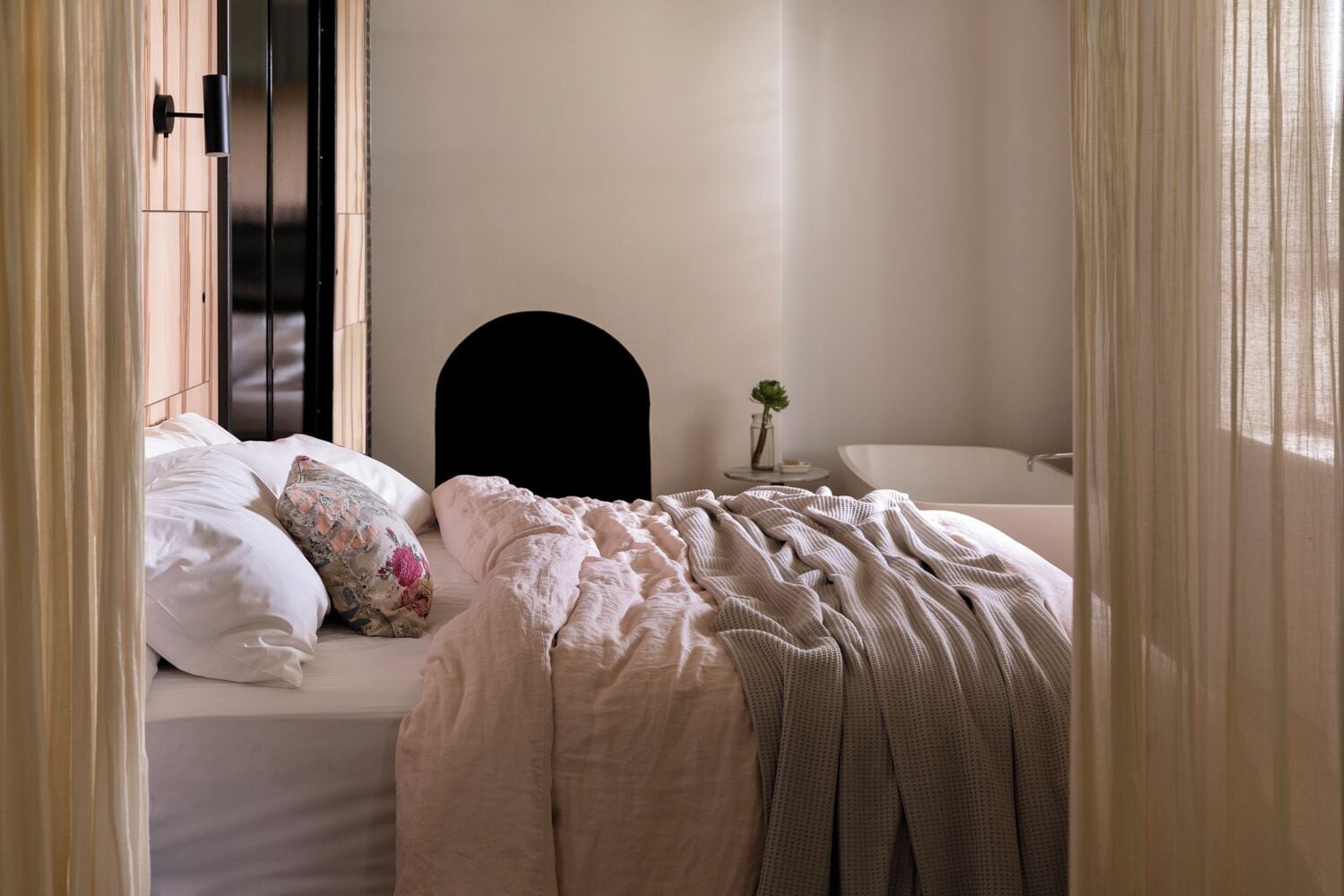 A bedroom with a bed, a nightstand, a small table with a plant, and a bathtub.