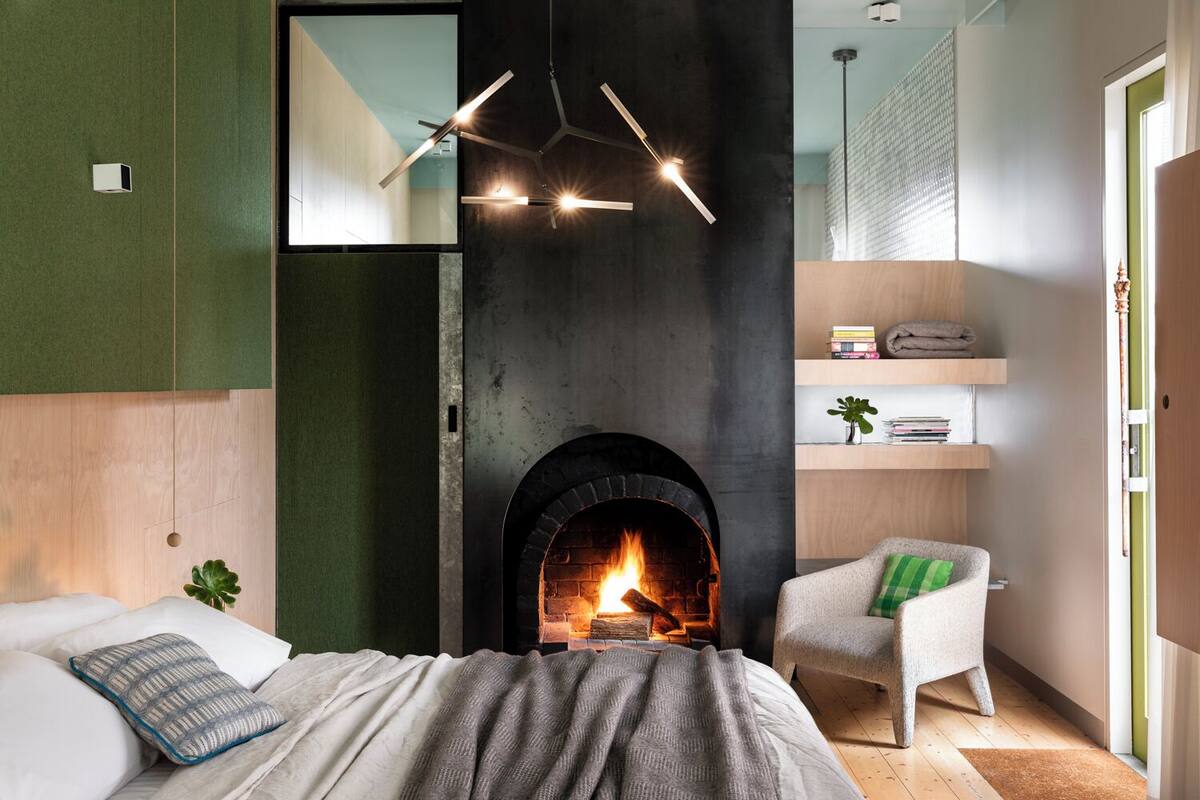 A modern bedroom with a fireplace, a bed with pillows, a chair, and a shelf with books and a plant.