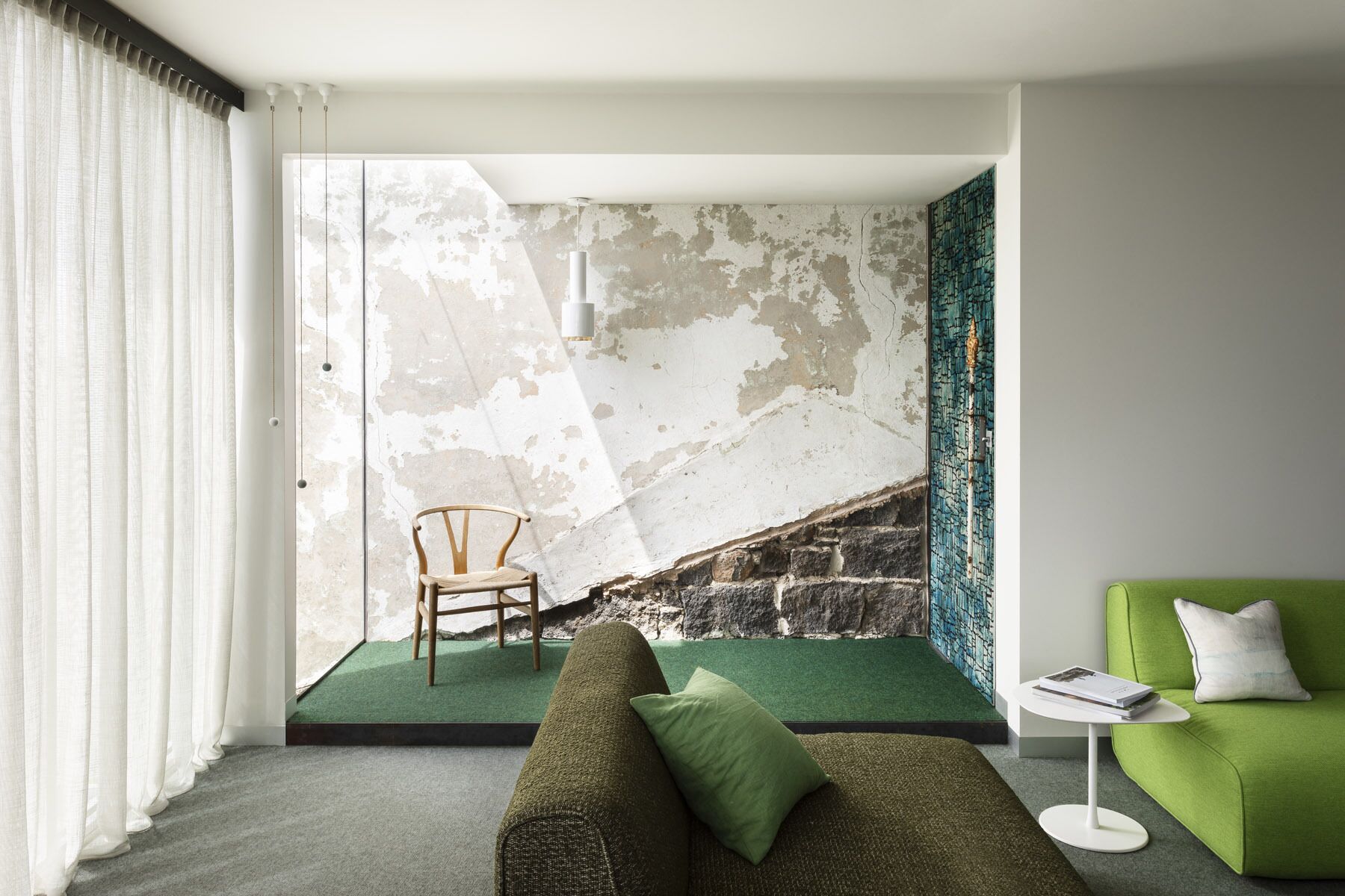 A modern living room with a green sofa, a green armchair, a wooden chair, and a small white table with a book.