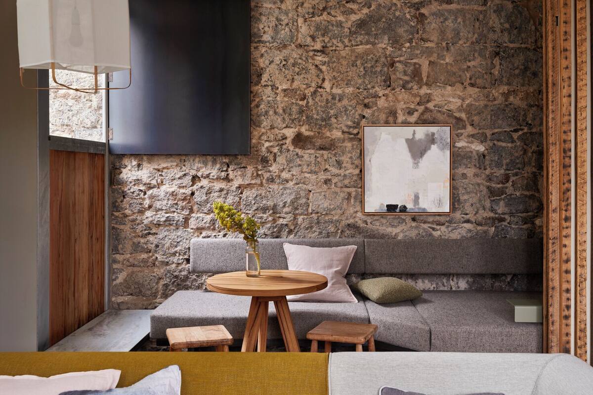 A cozy room with a stone wall, a grey sofa, a round wooden table with a vase, and a mounted artwork.