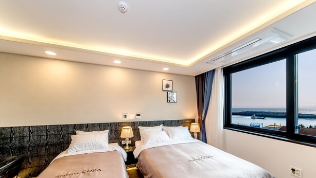 Habitación de hotel con dos camas, cabecero de madera, lámparas de noche, ventana con vista al mar y aire acondicionado.