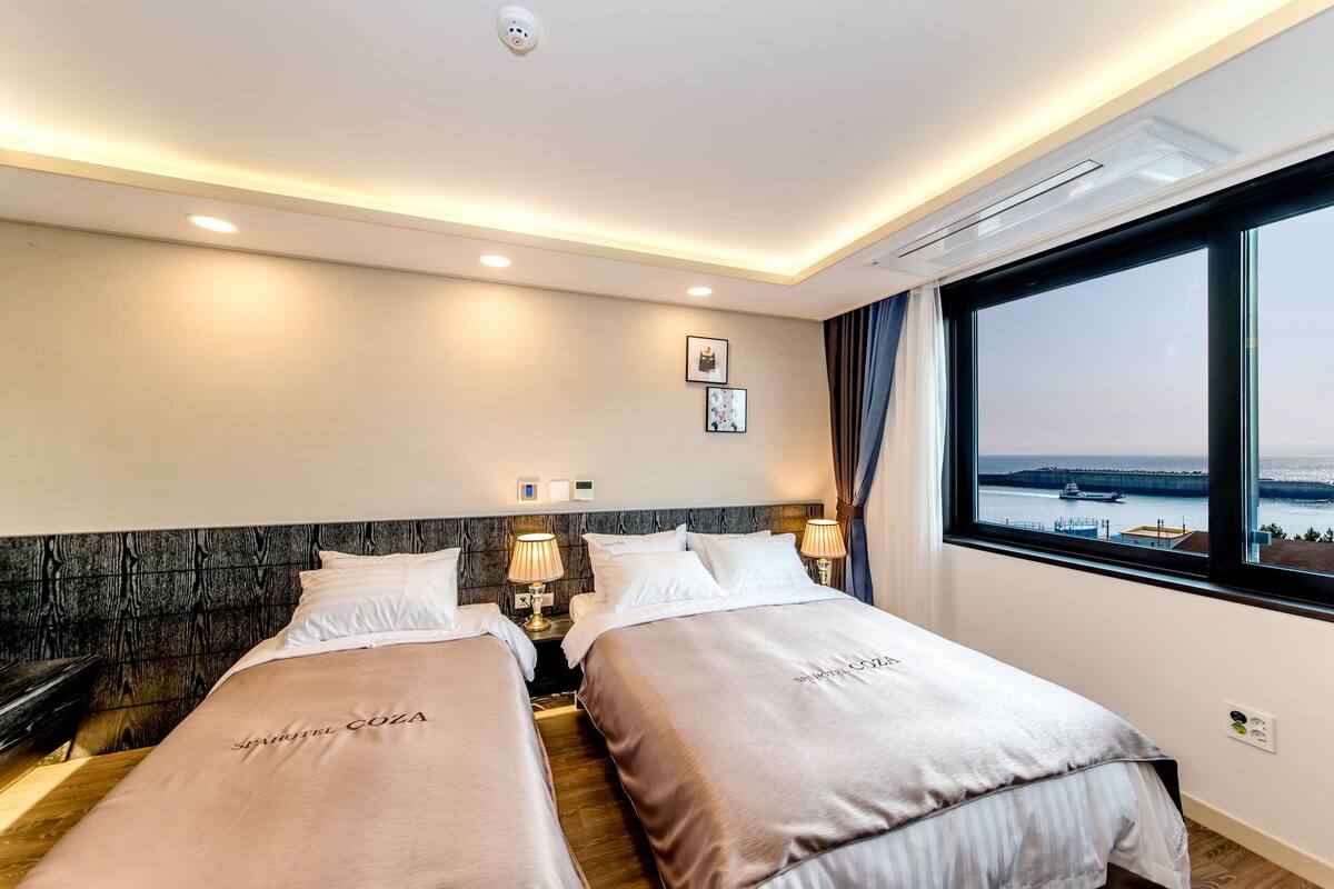 Een hotelkamer met twee bedden, een houten hoofdbord, bedlampen, een raam met uitzicht op zee en een airconditioner.