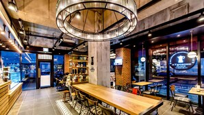 Un café moderno con una larga barra de madera, varias zonas de estar y una gran lámpara circular.