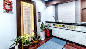 Una zona de recepción con un letrero que dice "SPA HOTEL COZA", un reloj rojo y plantas en macetas.