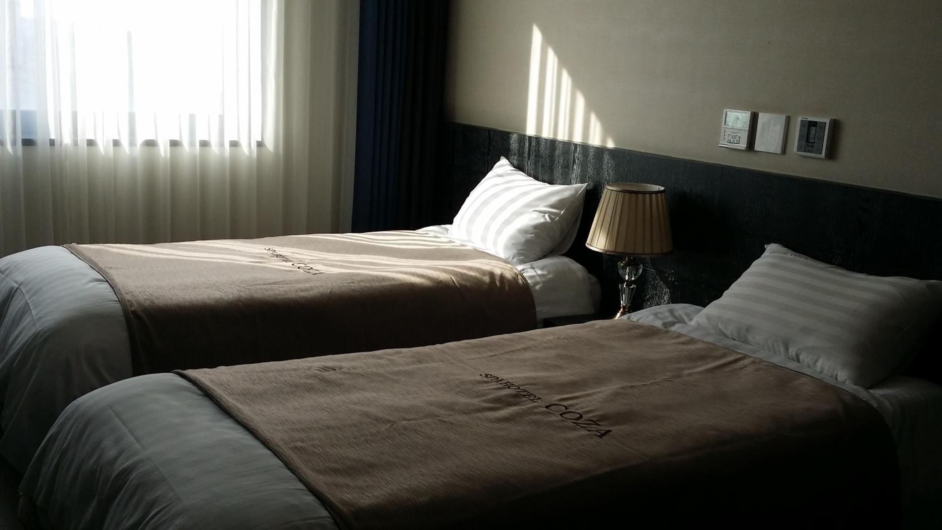 Habitación de hotel con dos camas, cada una con una colcha marrón y almohadas blancas. Hay una lámpara sobre una mesita de noche y un termostato en la pared.