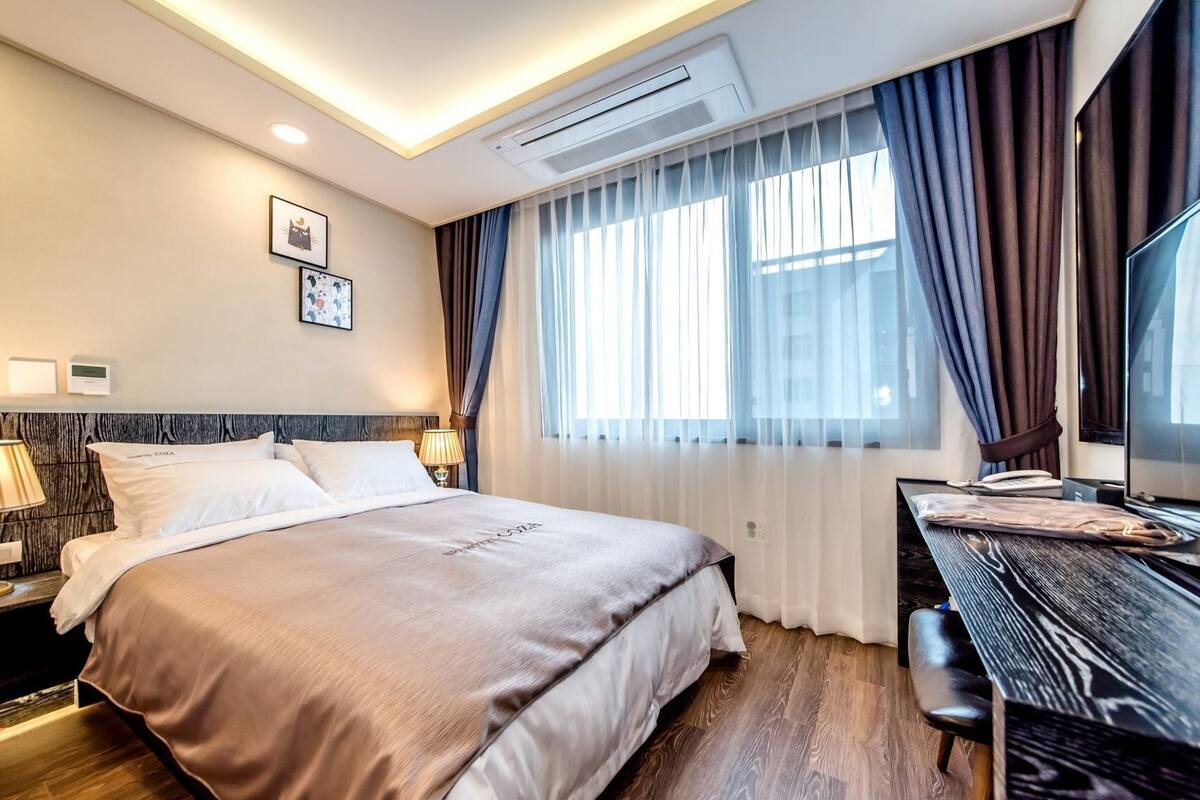 Een hotelkamer met een bed, nachtlampjes, een bureau met stoel, een televisie en een raam met gordijnen.