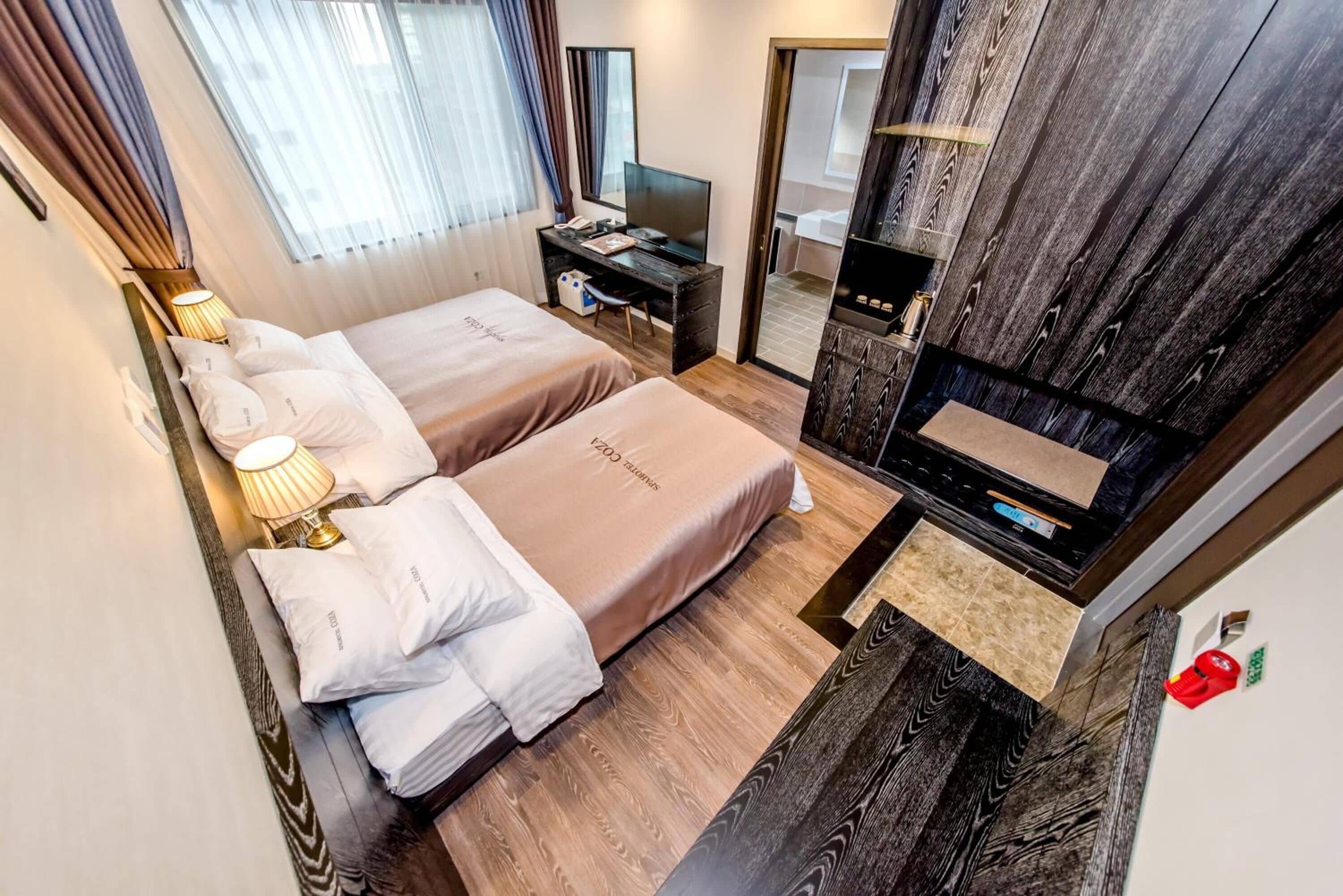A hotel room with two beds, a desk with a computer, a TV, and a bathroom visible through an open door.