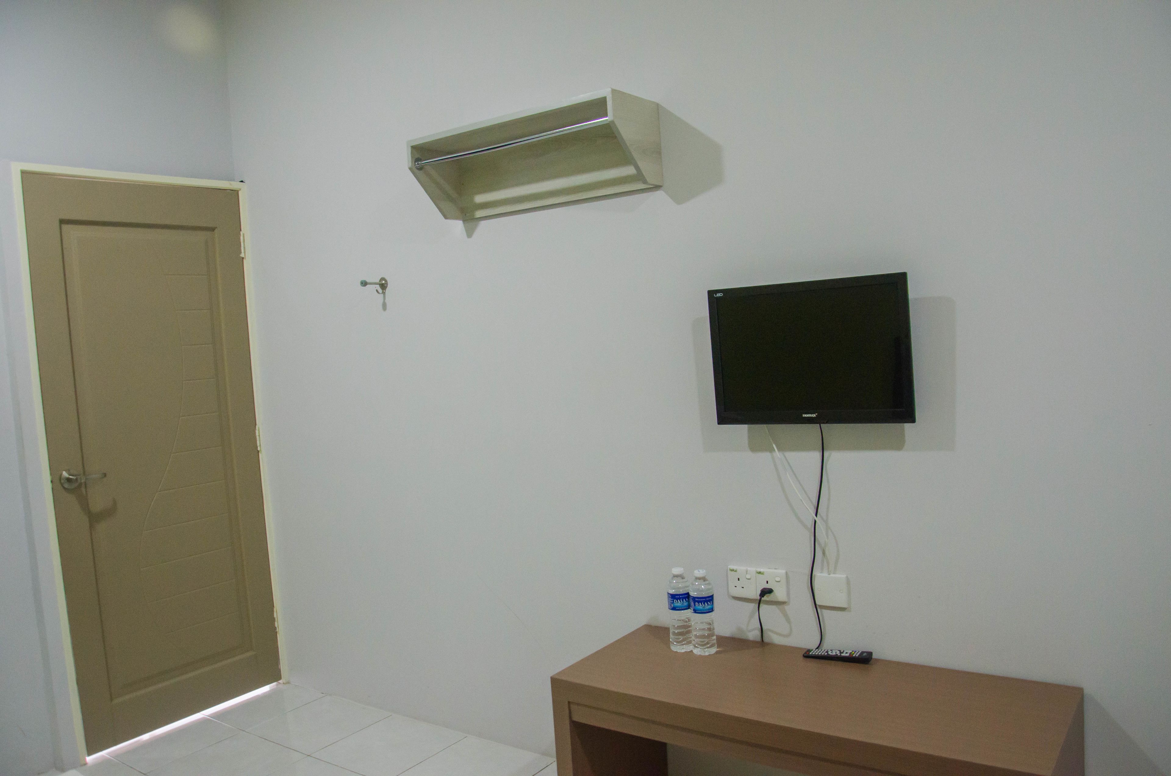 A room with a closed door, a mounted TV, a small table with two water bottles, and a light fixture on the wall.