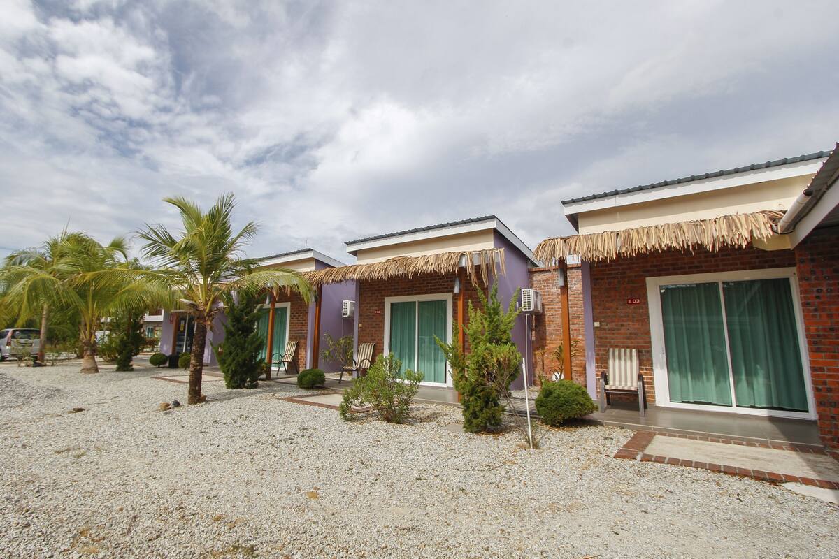 A row of two-story buildings with thatched roofs, a gravel driveway, and palm trees.