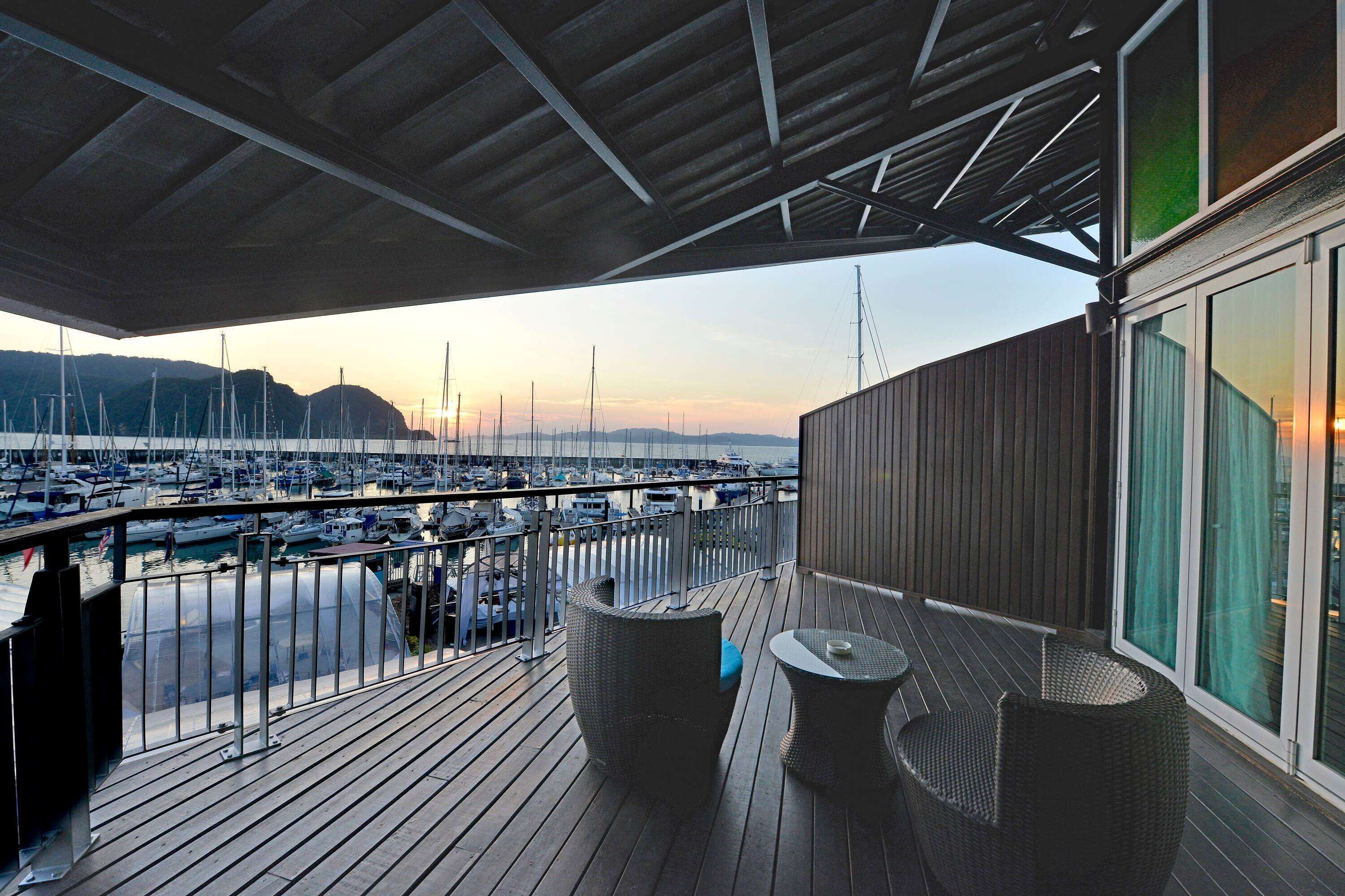 Un balcón con muebles de mimbre que da a un puerto deportivo con barcos y una puesta de sol al fondo.