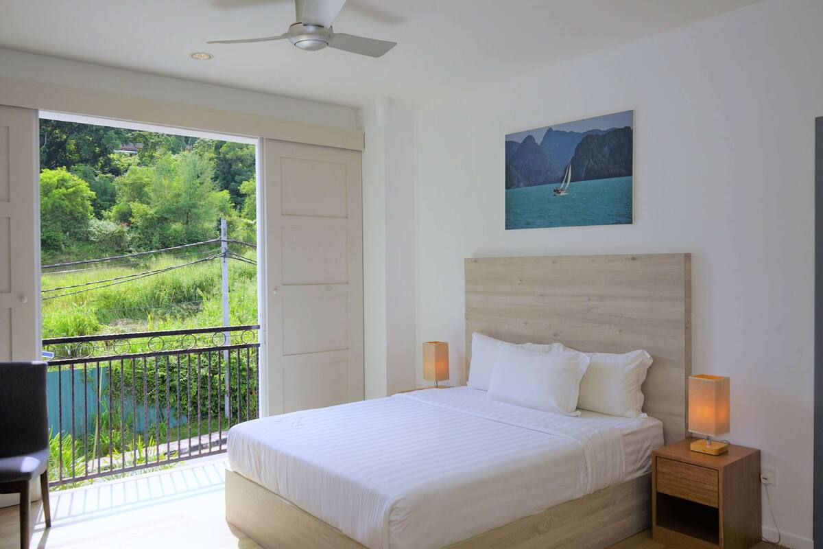 Un dormitorio con una cama grande, una mesita de noche con lámpara, un cuadro de una escena costera y un balcón con vistas a la vegetación y una casa distante.