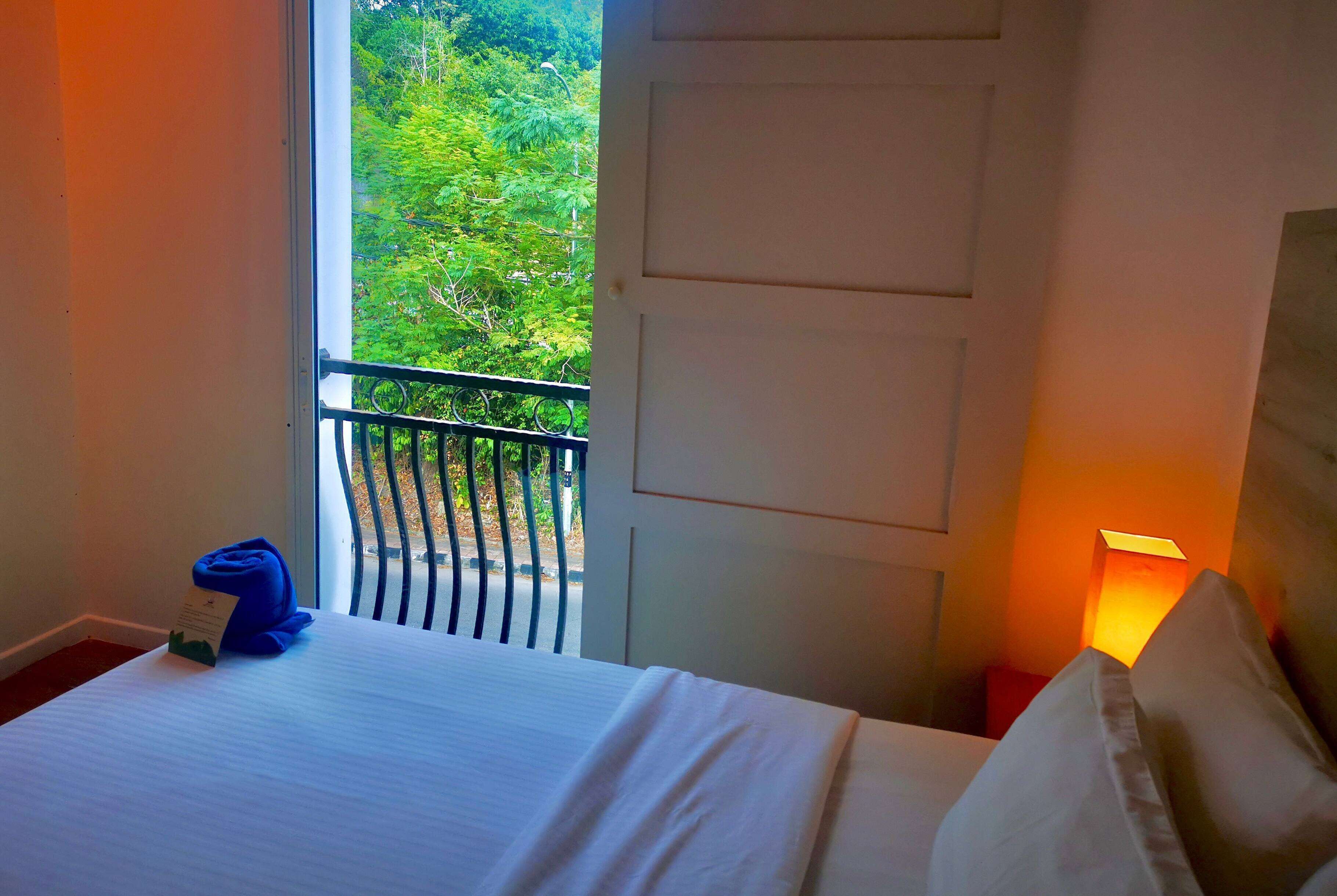 A hotel room with a bed, a nightstand with a lamp, a window with a view of greenery, and a door.