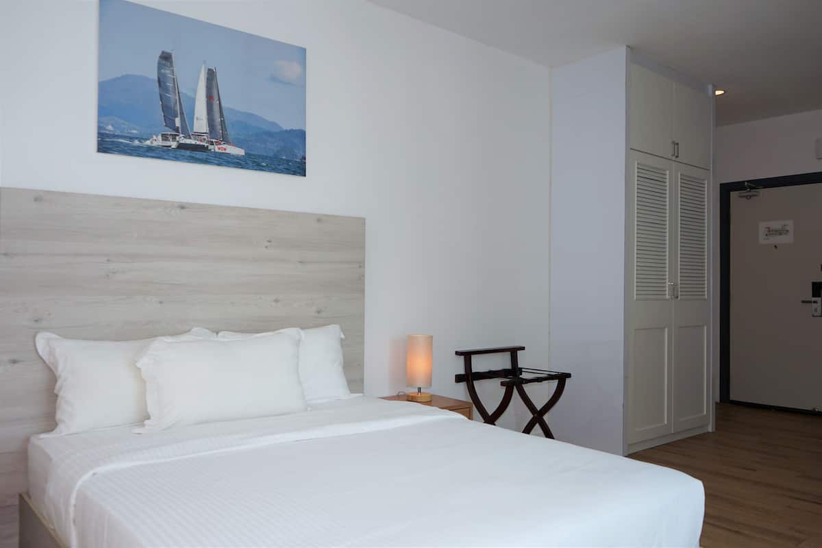 Une chambre d’hôtel comprenant un lit, une table de chevet, une lampe, un tableau représentant des voiliers et une armoire.