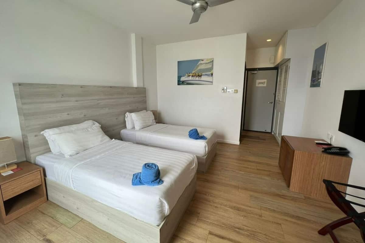 Habitación de hotel con dos camas, ventilador de techo, cabecero de madera, escritorio pequeño con silla, televisión y puerta que da a otra habitación.