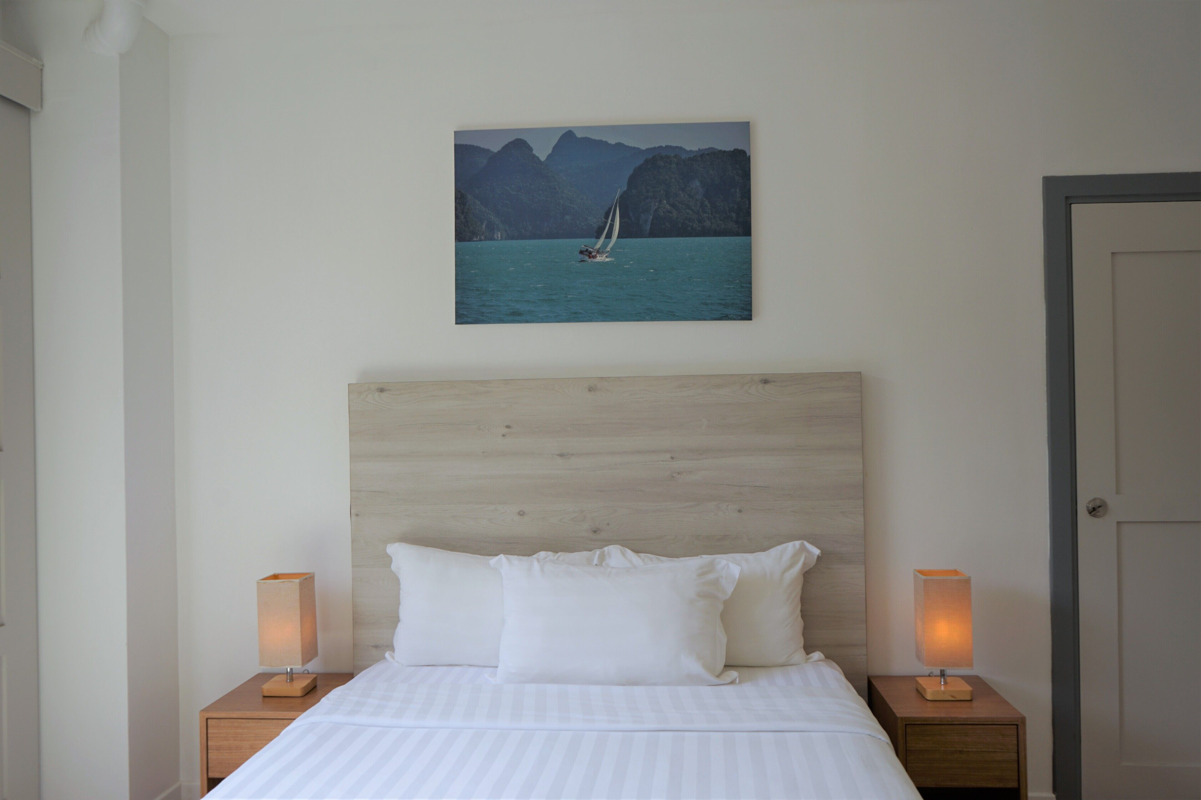 Un dormitorio con cabecera de madera, una cama con ropa de cama blanca, dos mesitas de noche con lámparas y un cuadro de un velero sobre la cama.