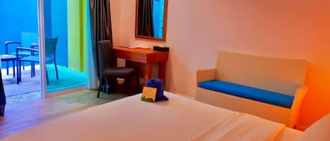 A hotel room with a bed, a desk, a chair, and a sofa. There is a sliding glass door leading to an outdoor area with a table and chairs.