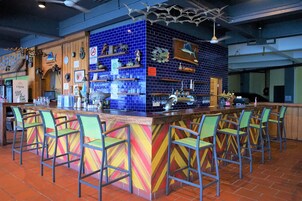 Una zona de bar con una pared de azulejos azules, estantes de madera con diversos objetos y sillas verdes.