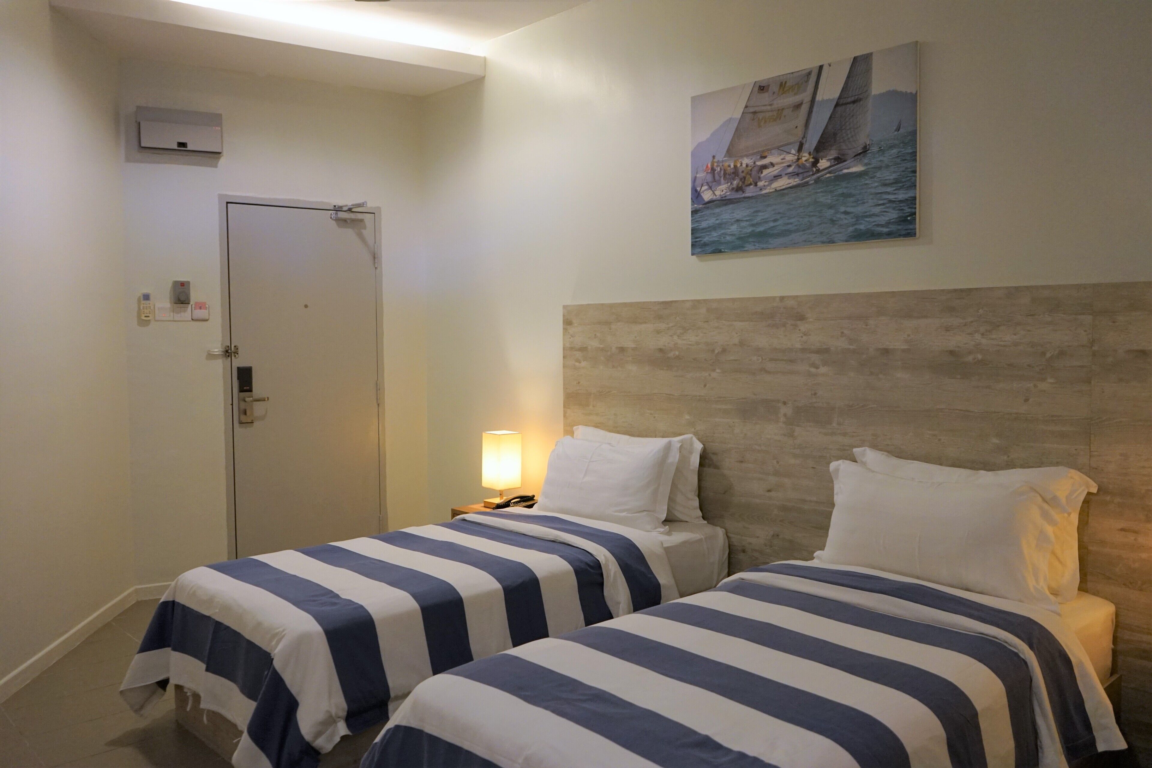 A hotel room with two beds, a wooden headboard, a painting of a sailboat, a door, and a nightstand with a lamp.