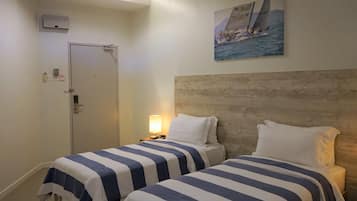 A hotel room with two beds, a wooden headboard, a painting of a sailboat, a door, and a nightstand with a lamp.