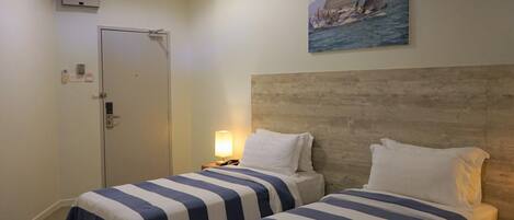 A hotel room with two beds, a wooden headboard, a painting of a sailboat, a door, and a nightstand with a lamp.
