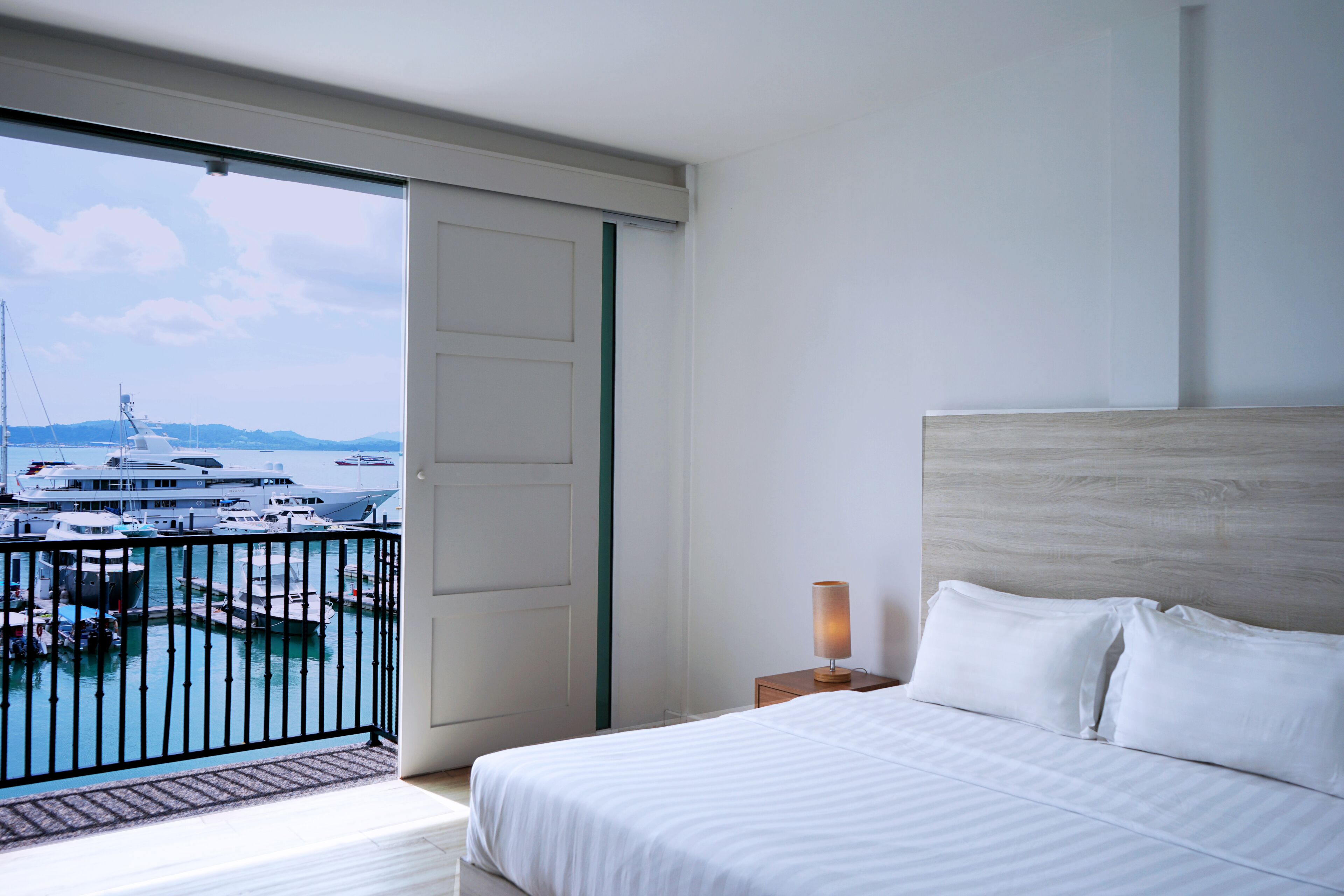 Habitación de hotel con una cama grande, una mesita de noche con lámpara y vistas a un puerto deportivo con barcos.